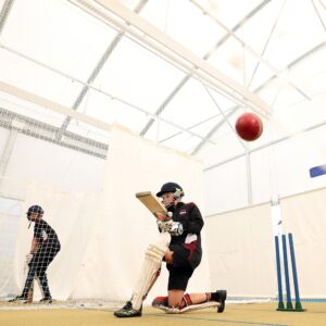 Young cricketer practising a batting shot in an indoor cricket net while a red cricket ball approaches the bat.