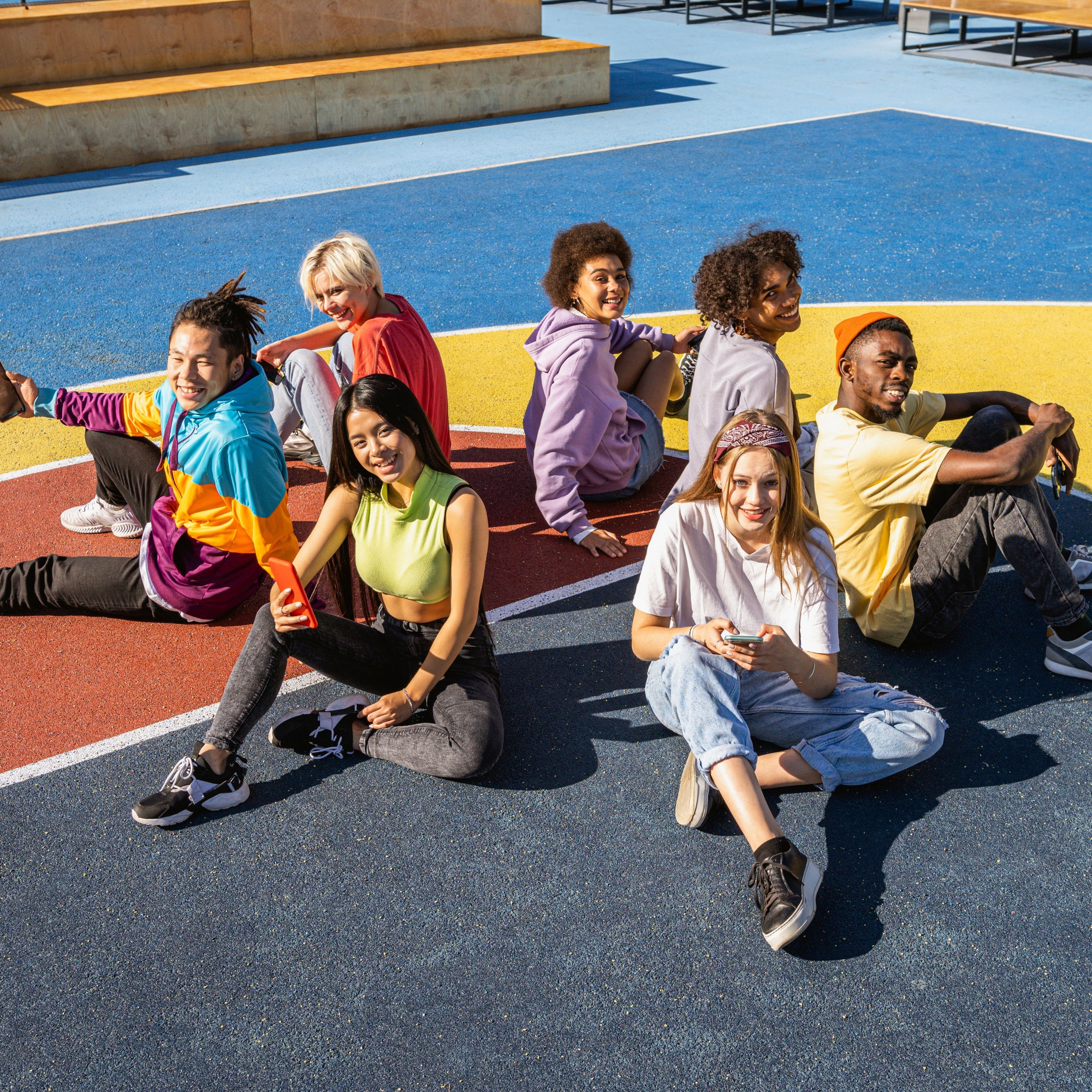 Diverse group of young people sitting together outdoors on a colourful sports court, smiling and socialising.